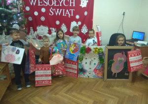Zdjęcie przedstawia dzieci stojących z prezentami pod napisem Wesołych Świąt